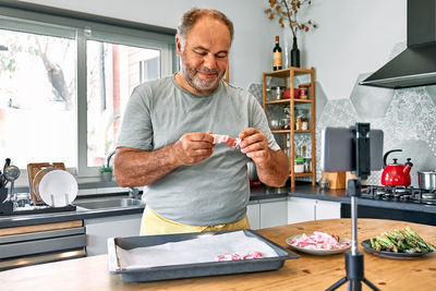 Man blogger preparing young green asparagus sprouts wrapped in bacon on wooden table in the kitchen.