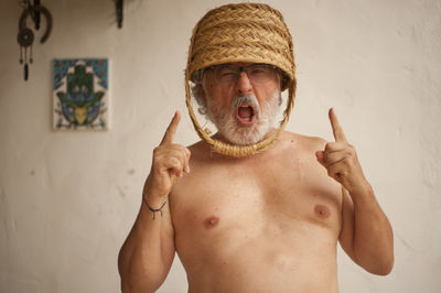 Portrait of shirtless man with basket on head shouting against wall at home