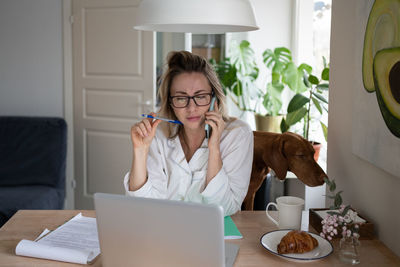 Woman with dog talking over phone by laptop on table
