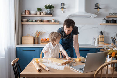 Cute preschool boy and his father baking cookies using video lesson on laptop