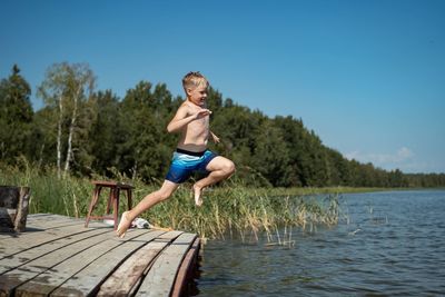 Cute caucasian boy jumping from wooden pier diving into lake in countryside