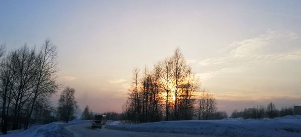 Bare trees on snow covered field against sky during sunset