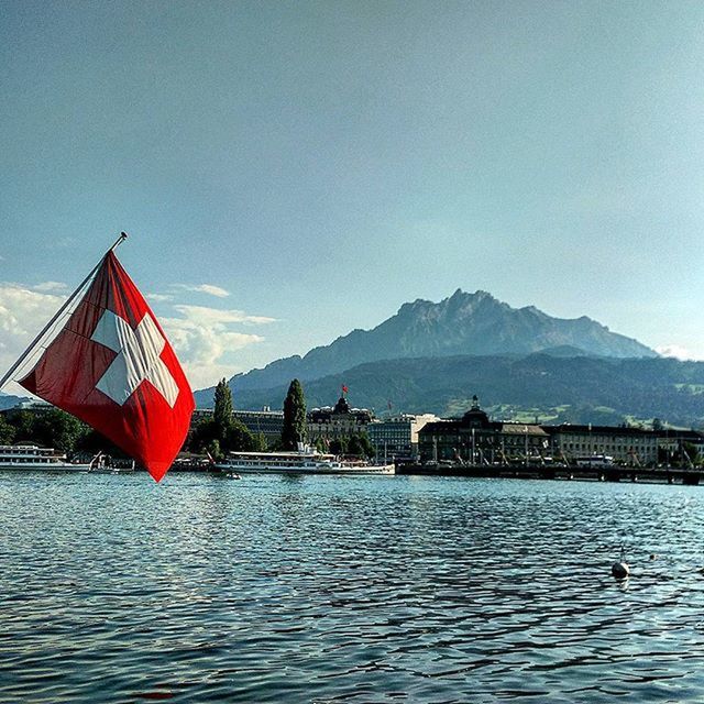 mountain, water, waterfront, nautical vessel, clear sky, flag, sky, lake, boat, transportation, sea, building exterior, blue, mountain range, built structure, architecture, rippled, tranquility, tranquil scene, copy space