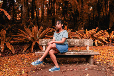 Full length of young woman sitting on bench in forest during autumn