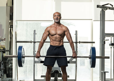 Portrait of shirtless man holding barbell standing in gym