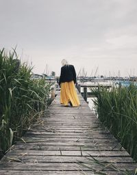 Rear view of woman on wooden walkway amidst plants against sky