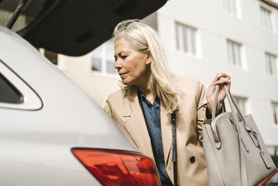 Low angle view of blond businesswoman holding purse while standing by car trunk