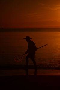 Silhouette man fishing in sea against orange sky