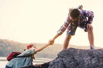 Boy helping girl climbing on rock against sky