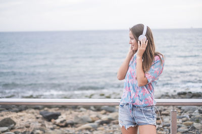 Young woman listening to music on headphones while standing against sea