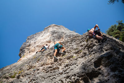 Low angle view of male friends climbing on mountain against clear blue sky