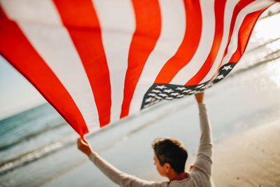 Rear view of man holding american flag while looking at sea