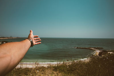 Close-up of cropped hand reaching towards sea against clear sky