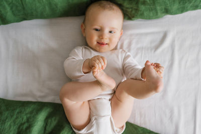 Top view of a charming smiling caucasian 6-month-old boy dressed in a bodysuit, holding his legs 