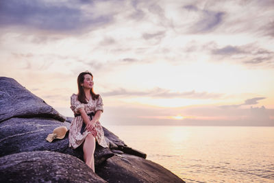 Woman sitting on rock by sea against sky during sunset