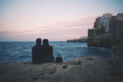 Rear view of couple sitting on rock by sea against sky during sunset