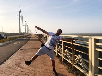 Playful man with arms outstretched standing on bridge against clear sky