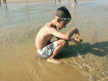 Shirtless boy playing with sand while crouching at beach