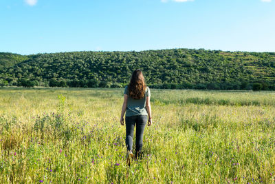 Rear view of young woman walking on grassy field against sky during sunny day