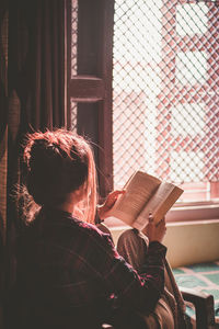 Woman reading book while sitting on bed by window