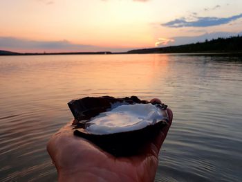 Person holding ice in lake against sky during sunset