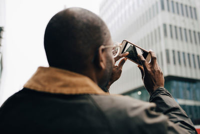 Rear view of businessman photographing modern building with smart phone in city