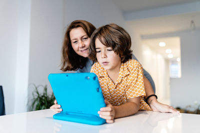 Cheerful kid with bright netbook at table near content mother in house with glowing lights