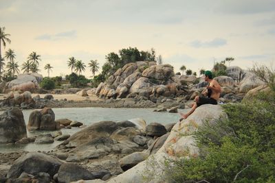 Side view of shirtless mid adult man playing guitar while sitting on rock at beach against sky during sunset