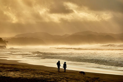 Silhouette peoplewalking at beach against sky during sunrise