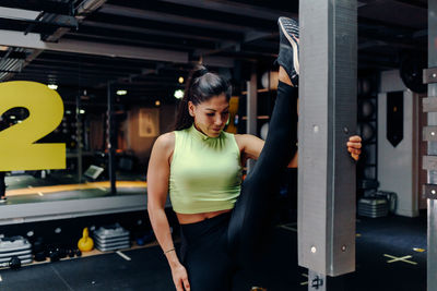 Full body young sporty female in fitness outfit performing stretching exercise near metal bar while warming up during training in gym