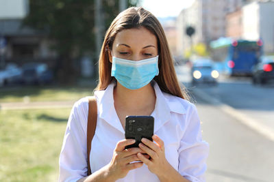 Woman wearing mask using mobile phone while standing outdoors