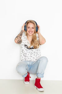 Portrait of smiling woman holding headphones while crouching against wall