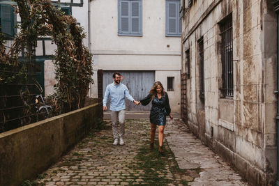 Cheerful young couple in stylish casual clothes holding hands and smiling while walking on old narrow street in city