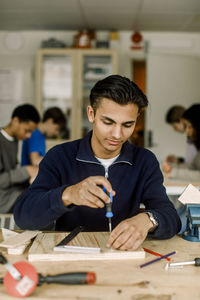 Male teenage student using screwdriver on wood while learning in carpentry class at high school