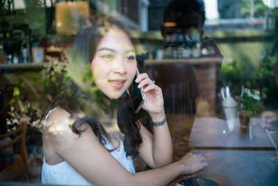 Woman using mobile phone while sitting at table seen through glass window