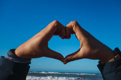 Cropped hands making heart shape at beach against clear blue sky