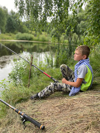 Boy sitting on fishing rod by lake