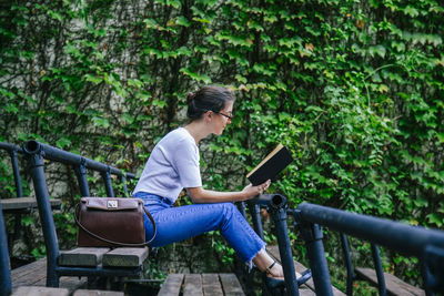 Full length of young woman using mobile phone while sitting on railing