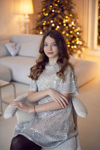 Teenage girl is sitting on a chair against the  of a christmas tree at home in a sequins dress