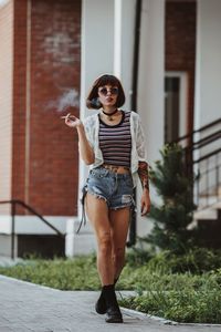 Full length of woman holding cigarette while walking on footpath