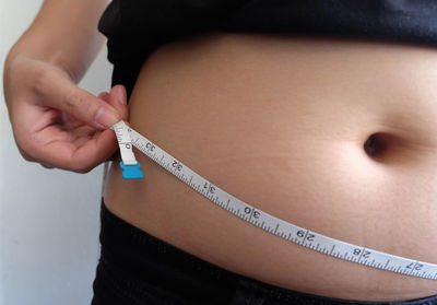 Midsection of overweight woman holding tape measure while standing against wall