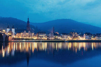 Illuminated buildings by lake against sky in city at dusk