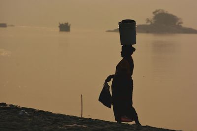 Silhouette woman with bucket on head walking at beach during sunset