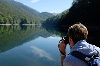 Rear view of woman photographing by lake against sky