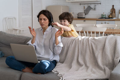 Exhausted mother sitting on couch at home, working on laptop, child distracted and asking attention