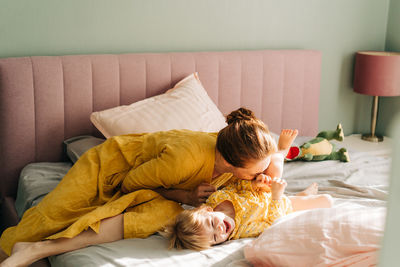 Mom tickles and kisses her little laughing daughter in the morning on the bed in the apartment.