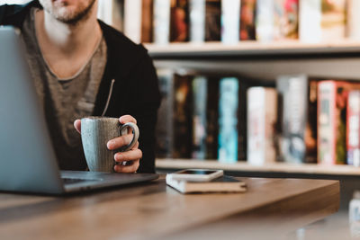 Midsection of man holding coffee cup while using laptop on table