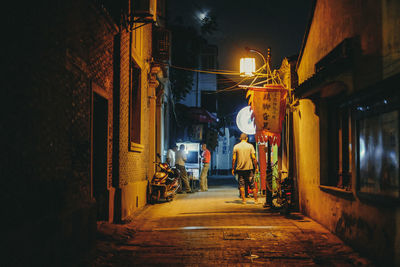 Rear view of man walking on illuminated street amidst buildings at night