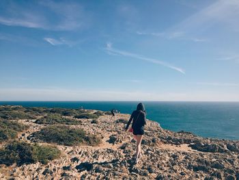 Rear view of woman walking rock formation by sea against sky