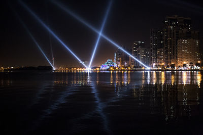 Illuminated buildings by river against sky at night